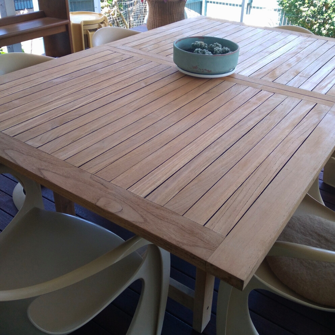 Wooden outdoor table with a green bowl on top, surrounded by white chairs. The teak is restored from grey using Semco Teak Cleaner.